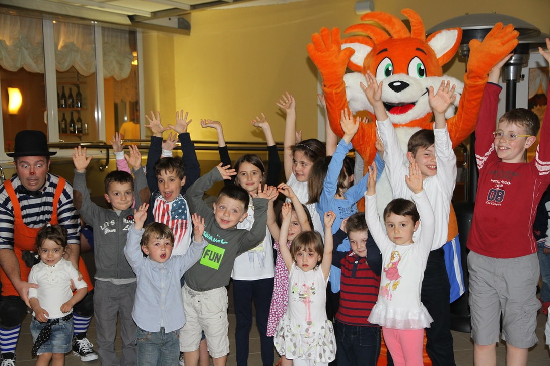 Eine Gruppe von Kindern mit erhobenen Armen nimmt an einer organisierten Unterhaltungsaktivität in einem Hotel teil. Sie werden von einem Animateur und demselben Fuchs-Maskottchen begleitet, was die Art der familienfreundlichen Aktivitäten während eines Hotelurlaubs zeigt.