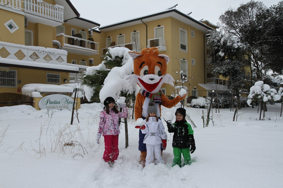 Eine Winterszene vor dem mit Schnee bedeckten Pinetina Mare Familienhotel. Drei Kinder in bunter Winterkleidung posieren mit dem Fuchs-Maskottchen. Dieses Bild vermittelt effektiv ein Familien-Winterurlaubserlebnis und zeigt sowohl die Unterkunft als auch Aktivitäten für Kinder.